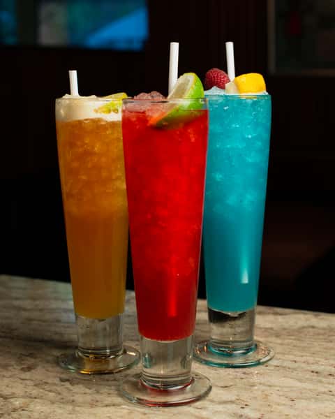 Colorful trio of iced cocktails—orange, red, and blue—garnished with fresh fruit and served in tall glasses on a marble bar in a trendy restaurant