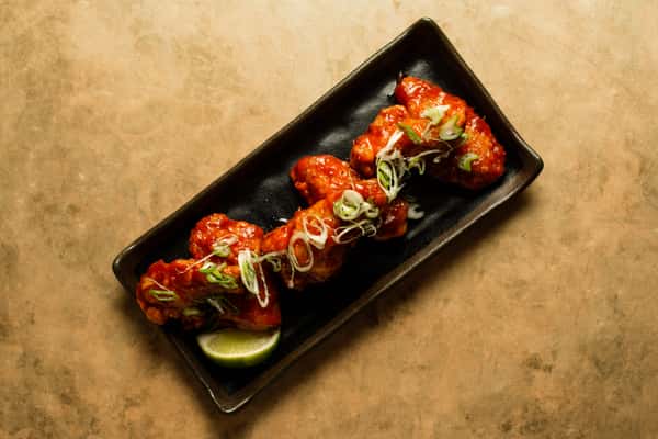 Crispy fried chicken wings coated in spicy sauce, garnished with scallions and lime, served on a sleek black plate in a modern Asian restaurant