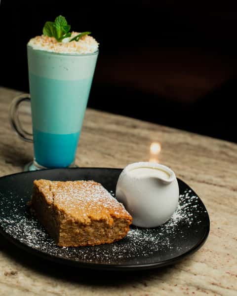 Moist slice of sponge cake dusted with powdered sugar, served with a side of cream and paired with a layered blue matcha latte topped with foam and mint in a cozy modern café setting