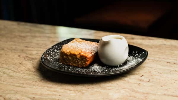 Moist slice of sponge cake dusted with powdered sugar, served with a side of cream on a modern black plate in a cozy café setting