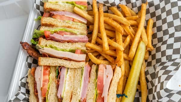 Club sandwich with ham, turkey, lettuce, tomato, and bacon, served with french fries and a pickle spear.
