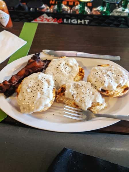 White plate with biscuits and sausage gravy, two strips of bacon, a fork, and a knife.