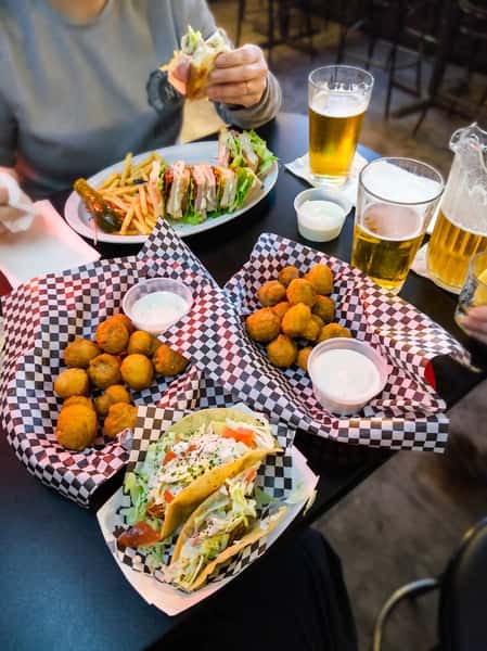 A table with beer glasses, a club sandwich with fries and a pickle, two baskets of fried mushrooms with ranch, and two tacos.