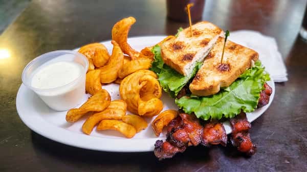 A sandwich on toasted bread with lettuce, bacon, curly fries, and a side of white dipping sauce on a white plate.