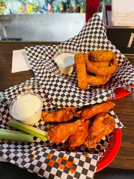 Buffalo wings with celery sticks and ranch dressing, plus a side of onion rings, served on checkered paper.