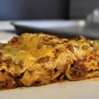 Close-up of a cheesy beef lasagna slice layered with seasoned ground meat, marinara, and gooey melted cheese on a white plate.