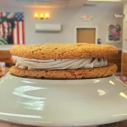 Soft cookie sandwich filled with a swirl of creamy vanilla frosting, presented on a white plate with a diner setting in the background.