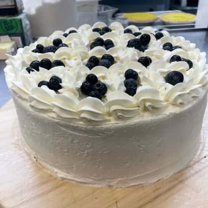 Whipped cream-frosted cake topped with fresh blueberries and piped rosettes, sitting on a wooden board in a kitchen prep area.