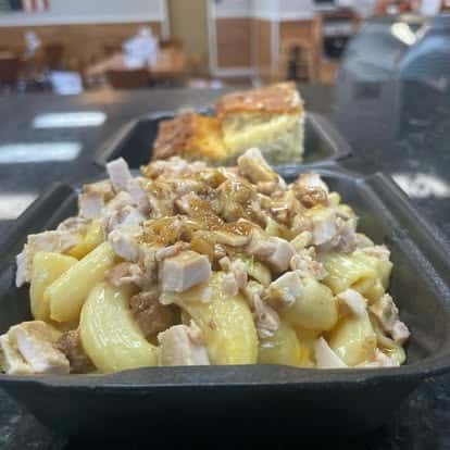 Macaroni pasta salad loaded with chunks of chicken, melted cheese, and savory seasoning, served in a black takeout tray with cornbread in the background.