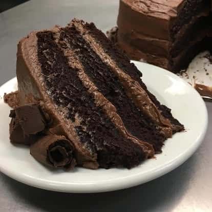 Rich slice of triple-layered chocolate cake with thick chocolate frosting and chocolate curls on a white dessert plate.