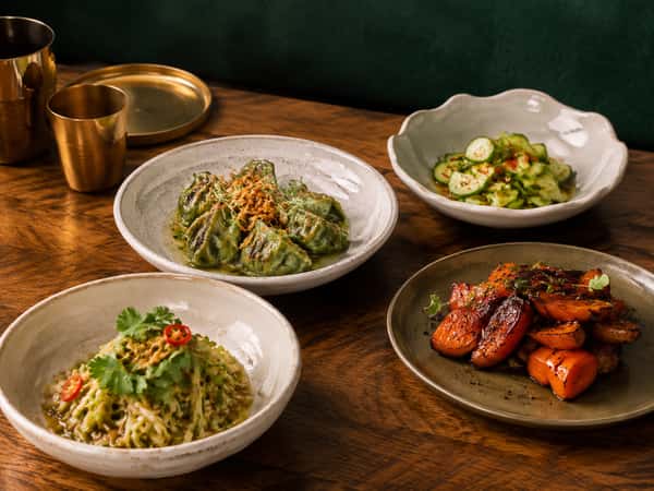 Assorted Asian-inspired restaurant dishes including green dumplings, spicy cucumber salad, fresh herb noodles, and glazed roasted vegetables served on a rustic wooden table.