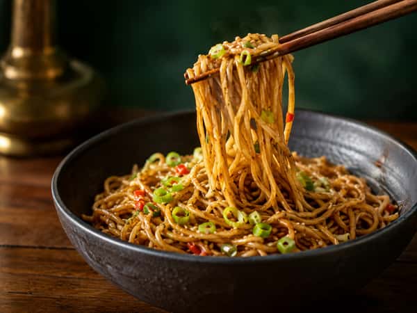 Chopsticks lifting savory stir-fried noodles with green onions and red peppers from a bowl at Tora Asian House
