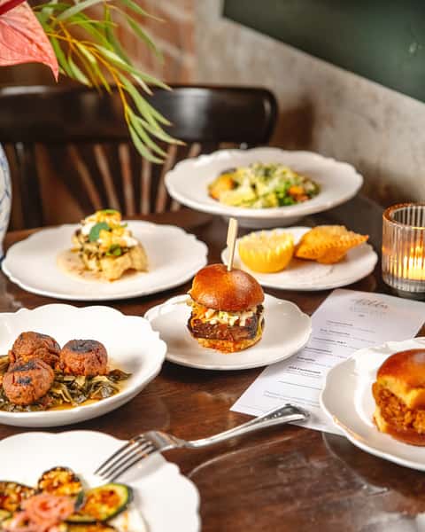 Assorted small plates with sliders, empanadas, grilled vegetables, meatballs, and pasta on a candlelit wooden table.