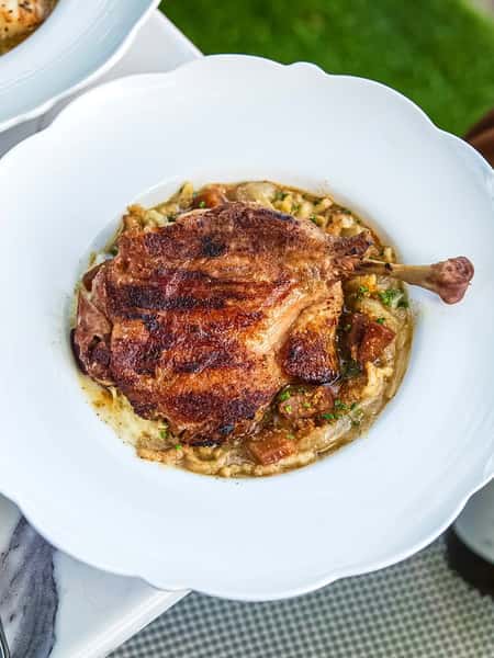 Crispy duck leg confit served over a creamy, rustic vegetable and herb ragout in a white scalloped bowl.