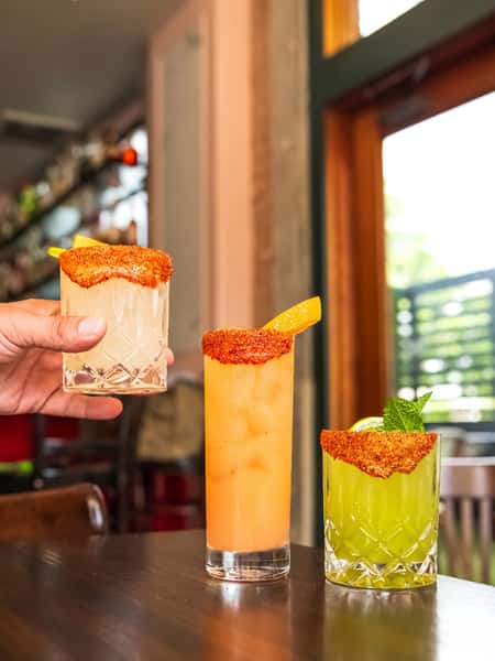 Trio of colorful cocktails with chili-lime rims, garnished with citrus and mint, served on a wooden table in a bright bar setting.