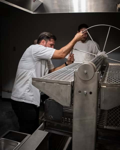 Chefs in white uniforms operating a large commercial rotisserie grill in a professional restaurant kitchen, showcasing behind-the-scenes culinary preparation with industrial-grade cooking equipment.
