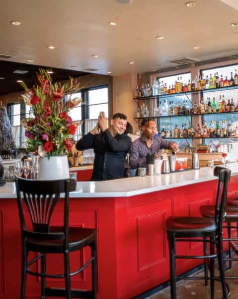 Bartenders crafting cocktails behind a vibrant red bar at a lively upscale restaurant, with a backdrop of neatly arranged premium spirits and fresh floral décor enhancing the welcoming ambiance.