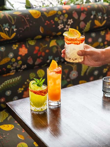 Three vibrant cocktails with tajín-rimmed glasses, garnished with citrus and mint, on a wooden table with floral booth seating.