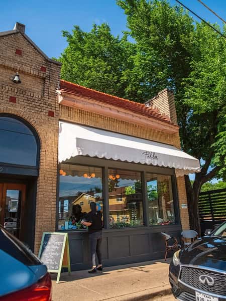 Street view of Pillar Bistro with white awning, large windows, sidewalk chalkboard menu, and a guest at the window.