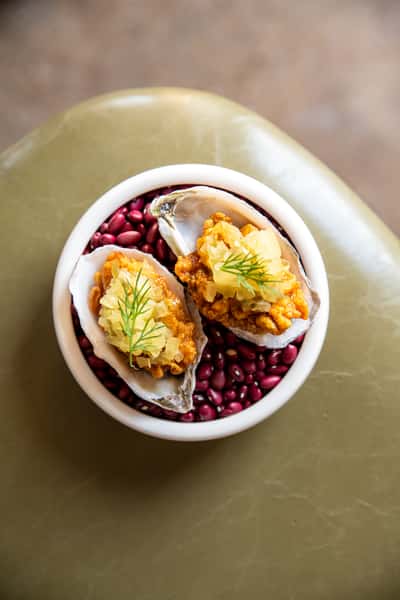 Two crispy fried oysters topped with pickled onions and fresh dill, elegantly served in their shells on a bed of red beans, presented in a white bowl on a green ceramic surface.