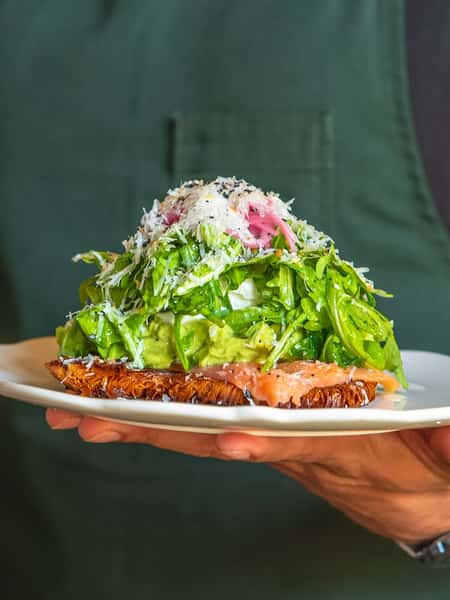 Open-faced smoked salmon and avocado toast topped with a poached egg, shredded greens, pickled onions, and grated cheese, served on a white plate—fresh brunch dish from a modern bistro.