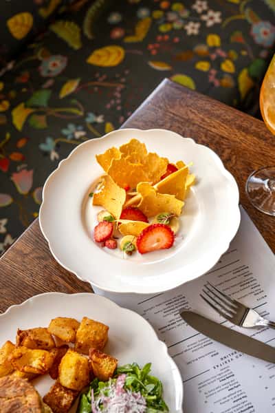 Strawberries with whipped cream, garnished with golden tuile crisps and microgreens on a scalloped white plate.