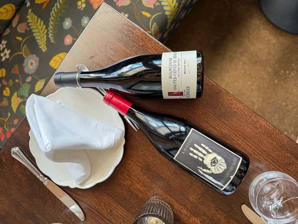 Two bottles of red wine rest on a wooden table set with a napkin-wrapped plate, knife, fork, and water glass at a cozy restaurant.