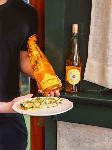 Server presenting Cà del Bosco sparkling wine with cucumber-topped toast; a second wine bottle displayed on a nearby ledge.