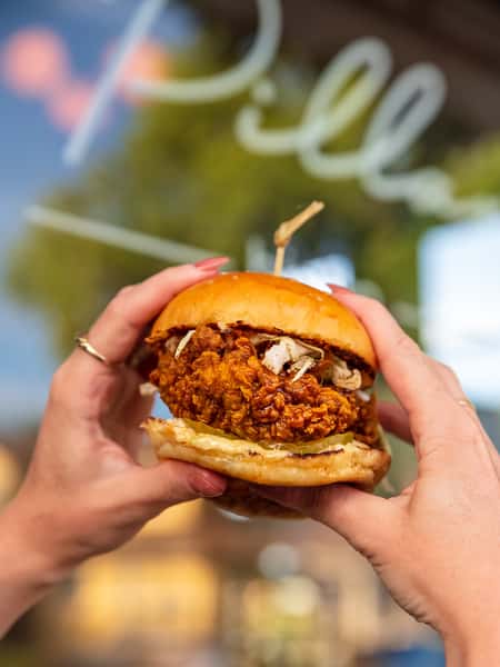 Crispy fried chicken sandwich with pickles and slaw on a toasted brioche bun, held up in front of a restaurant window.