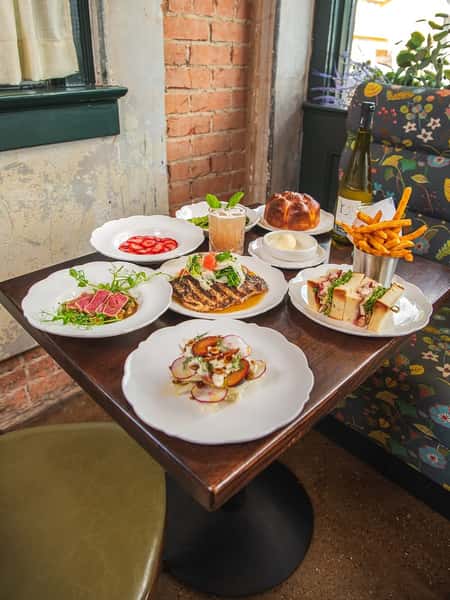 Vibrant table spread at a rustic-chic restaurant featuring seared tuna salad, grilled fish with citrus bean salad, fresh radish and burrata salad, strawberry-topped dessert, a stacked sandwich with fries, soft buttered roll with butter, and a mint-garnished cocktail, all paired with a chilled bottle of white wine.