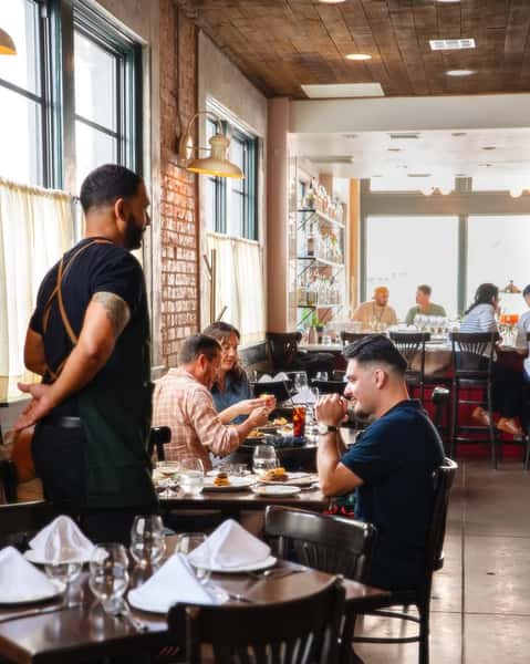 Lively interior of a trendy, upscale restaurant with exposed brick walls and large windows, featuring a bustling brunch scene with diners enjoying cocktails and gourmet dishes, attentive service from staff in modern aprons, and a cozy yet stylish ambiance. Ask ChatGPT