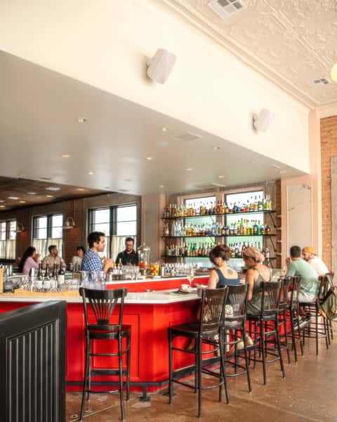Bright and modern bar with a bold red counter, high black chairs, and a wall of backlit liquor shelves, bustling with seated patrons.