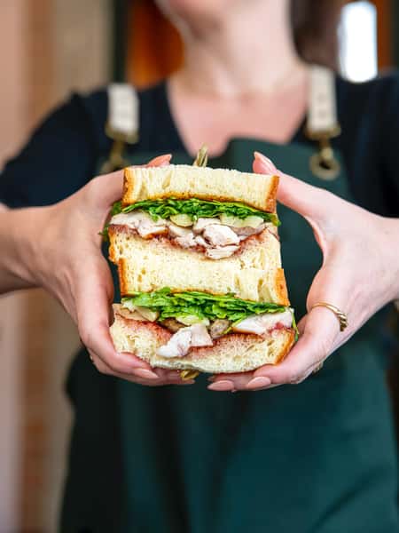 Stacked chicken sandwich on fluffy white bread with crispy greens and savory spread, held by a server in a green apron.