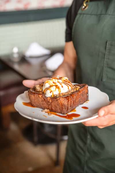 Thick-cut brioche French toast served with a generous dollop of whipped cream, drizzled with rich caramel sauce and sprinkled with crunchy granola, presented on a white scalloped plate at a cozy bistro.