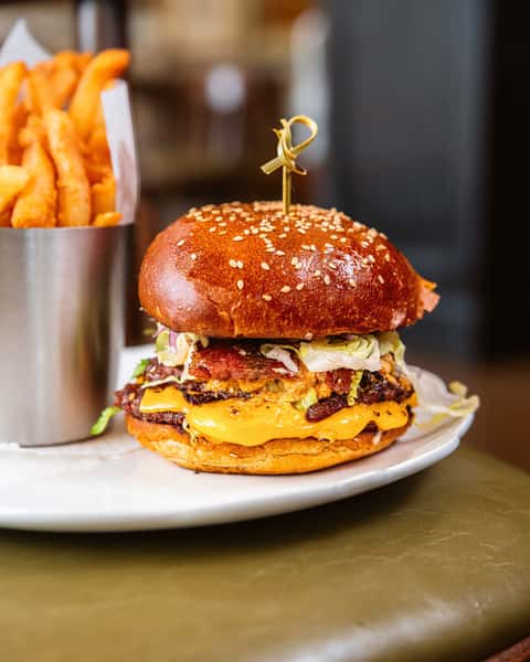 Juicy double cheeseburger with lettuce and bacon on a sesame brioche bun, served with golden fries in a metal cup.