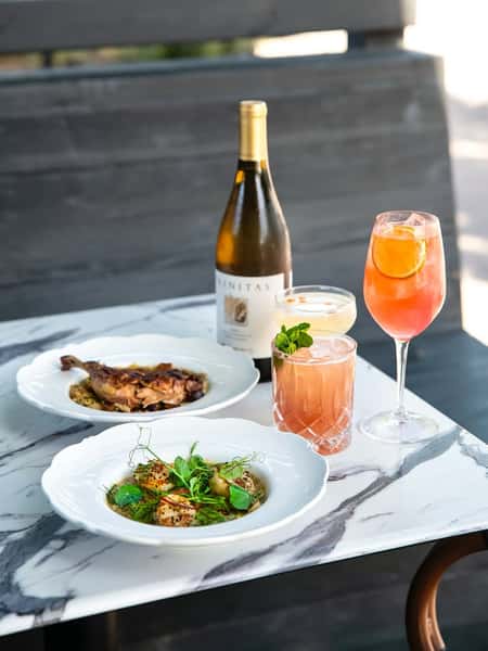 Elegant outdoor table setting with herb-crusted scallops, braised duck leg, cocktails, and a bottle of white wine.