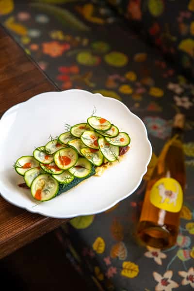 Toasted tartine topped with thin cucumber slices, dill, and hot sauce, paired with a bottle of chilled natural wine.