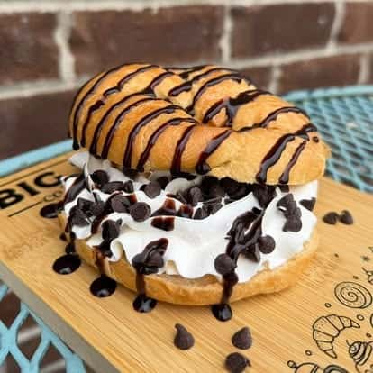 Croissant donut sandwich filled with whipped cream and chocolate chips, drizzled with chocolate syrup