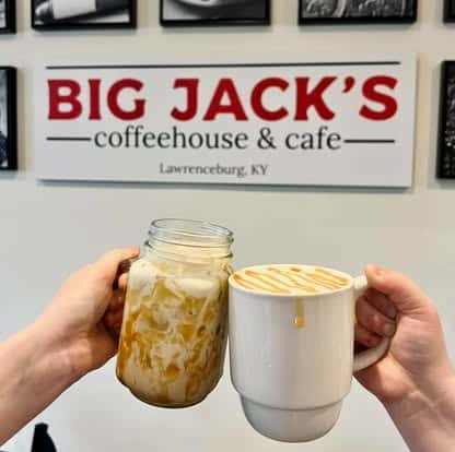 Two hands holding iced coffee drinks in front of a Big Jack's Coffeehouse & Cafe sign