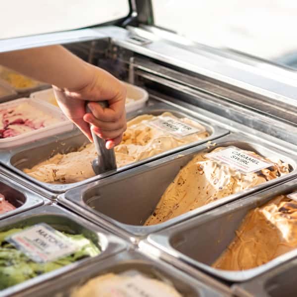 Hand scooping creamy gelato from a display case featuring caramel swirl and other artisan flavors