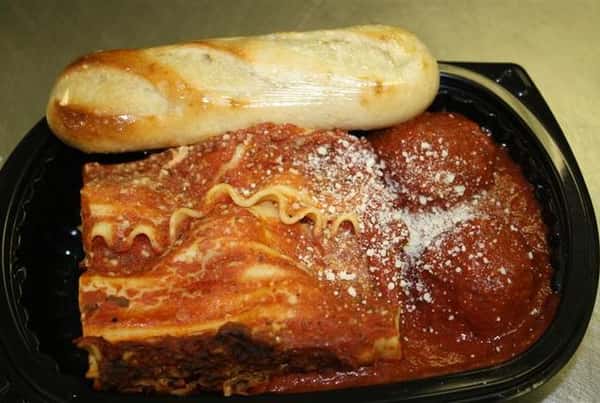 Takeout container with a piece of lasagna, two meatballs topped with parmesan cheese and a side of bread