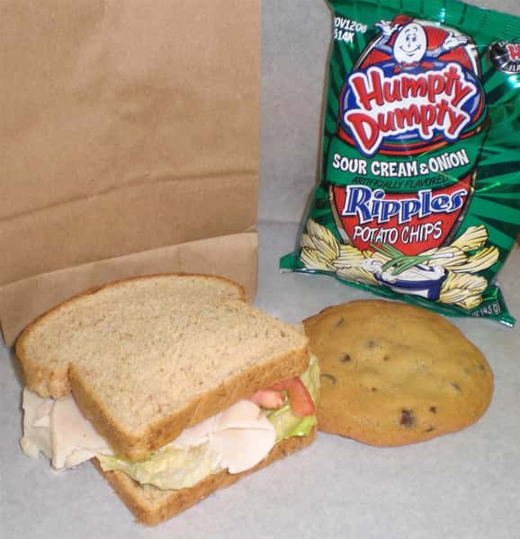 Sandwich with cheese, deli meat, lettuce and tomato with a side of sour cream and onion chips with a chocolate chip cookie