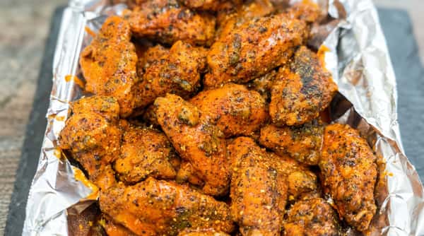 Close-up of orange-glazed chicken wings with visible herbs and black pepper, served in a foil container.