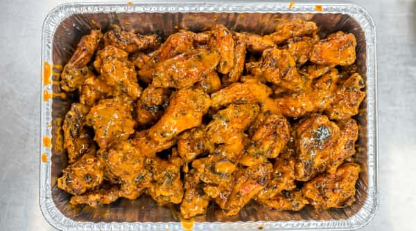 Buffalo chicken wings coated in orange sauce with visible herbs, served in a foil tray.