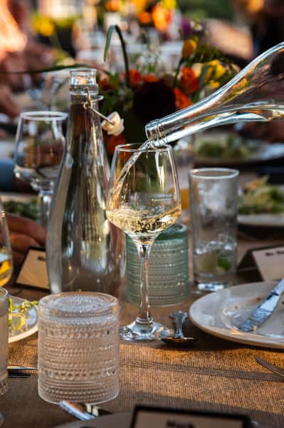 Crisp white wine being poured into a glass on a table that has been decorated for a private event. There are quirky glass cups, flower arrangements, and a pastel fabric table cloth.