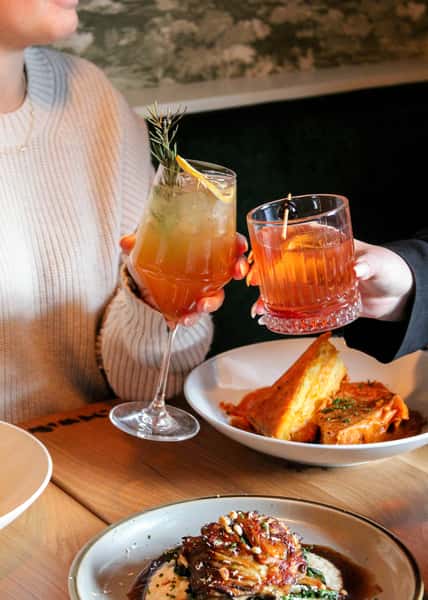 Two glasses contains red and orange liquid clink above a plate of grilled cheese