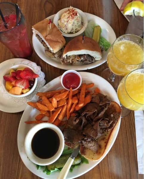 Two plates with steak sandwiches and a side of au jus, sweet potato fried and assorted drinks