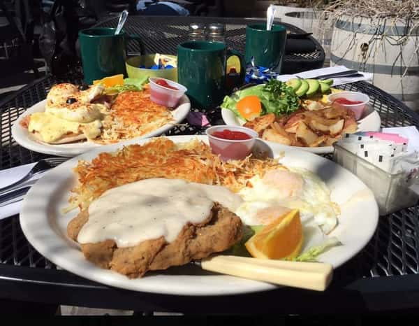 Chicken fried steak with two fried eggs and hasbrowns on a plate with two breakfast plates with assorted breakfast iems in the background