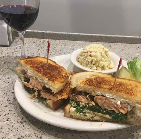 Seasoned salmon sandwich on toast bread with a side of potato salad and a glass of wine
