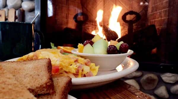 Omelet on a plate witha bowl of mixed fruit and slices of toast with a fire place in the background